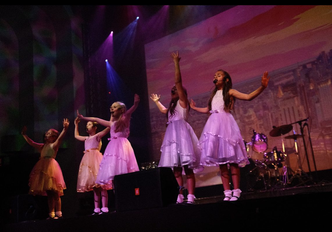 A group of young girls in purple dresses singing at a Bakerstown performance event